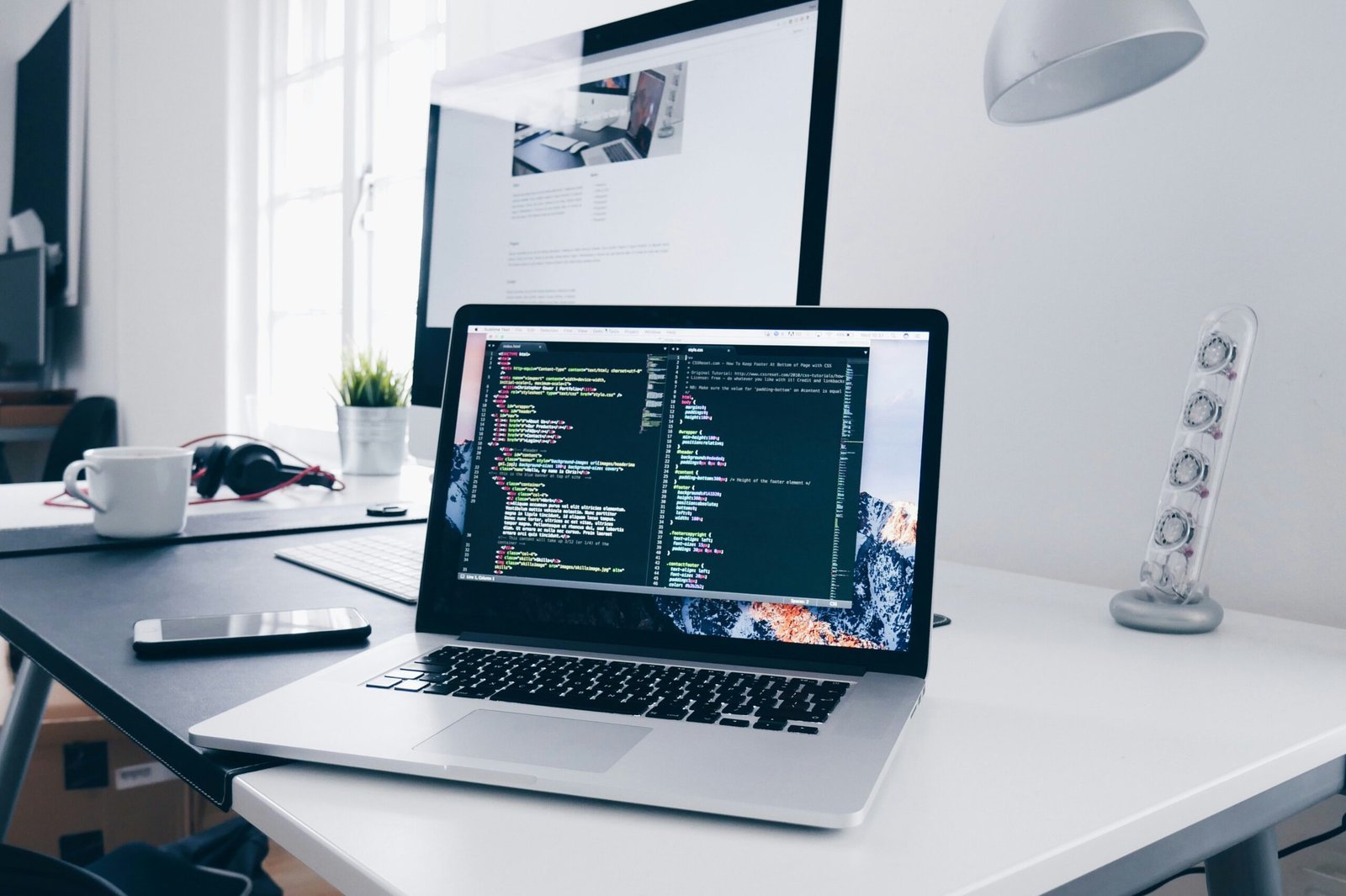 Heller Arbeitsplatz mit Laptop und Monitor, auf beiden ist Quellcode geöffnet